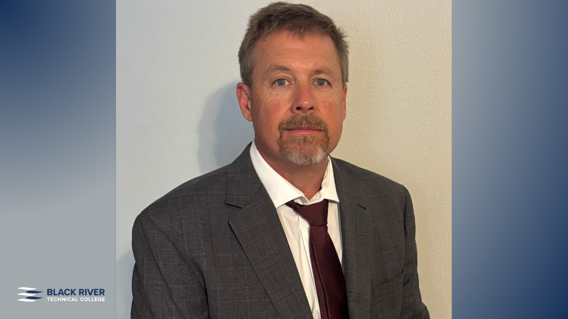 A man with short brown hair and a mustache, wearing a gray plaid suit and maroon tie, poses for a portrait against a plain wall, with Black River Technical College branding.